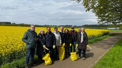 Die Teilnehmer des Fotospaziergangs, ausgestattet mit gelben Handschuhen und Müllsäcken. Im Hintergrund ist ein gelbes Rapsfeld zu sehen. 
